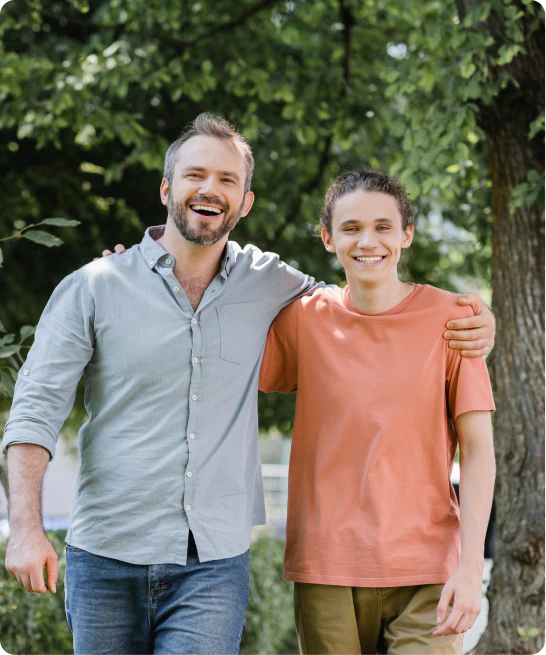 Two people standing outdoors under a tree, smiling at the camera. One is wearing a light blue shirt and jeans, the other is in a salmon-colored t-shirt and khaki pants. They have their arms around each others shoulders.