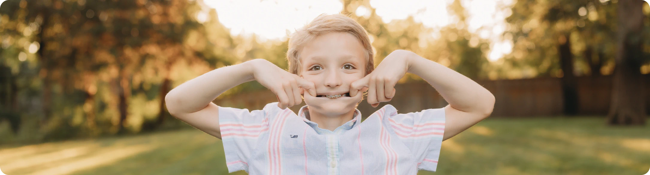 A young boy with blonde hair stands outdoors, smiling and pulling the corners of his mouth wide with both hands. He is wearing a short-sleeved, light-colored shirt with pink stripes on the sleeves. The background is a sunny, blurred yard.