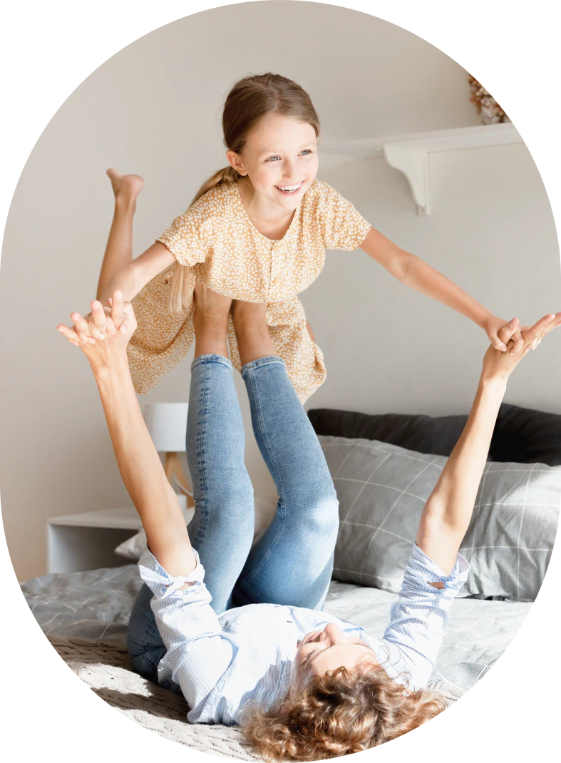 A woman lies on her back on a bed, holding a smiling young girl up in the air with her legs while they hold hands, playing together in a bright, cozy room.