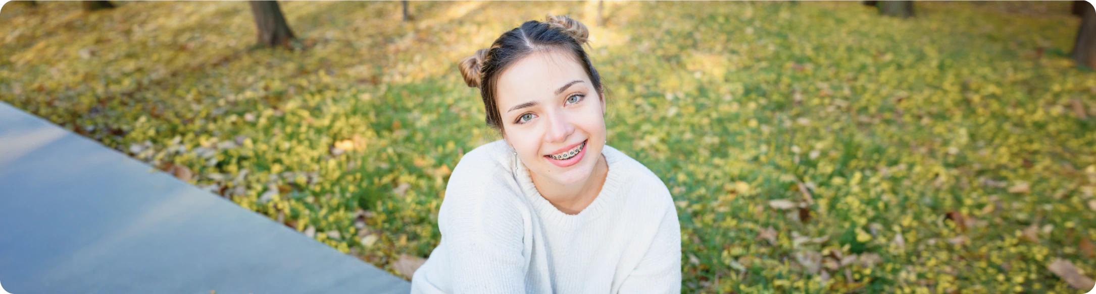 A young person with braces, wearing a white sweater and space buns, smiles while sitting outdoors on a grassy area covered with autumn leaves. Trees and a sidewalk are visible in the background.