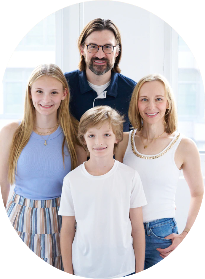 Mobile_MTD_Benjamin_Allen A family of four poses indoors, smiling. An adult man and woman stand in the back, and a girl and younger boy stand in front. They are dressed in casual, light-colored clothes with a bright window in the background.