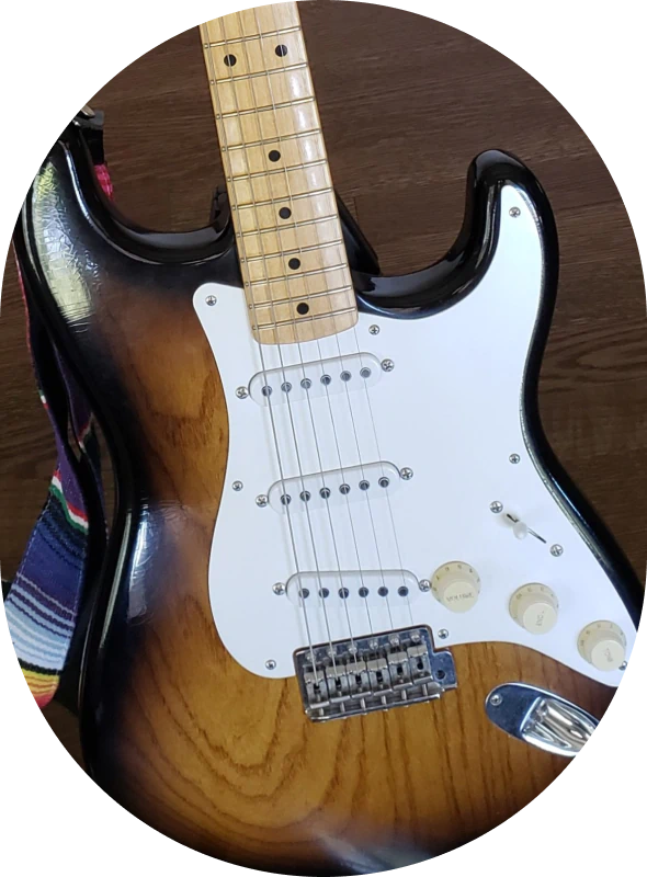 MTD_Beyond_The A close-up of an electric guitar with a sunburst finish, white pickguard, three single-coil pickups, and a colorful striped guitar strap, resting on a dark wooden surface.
