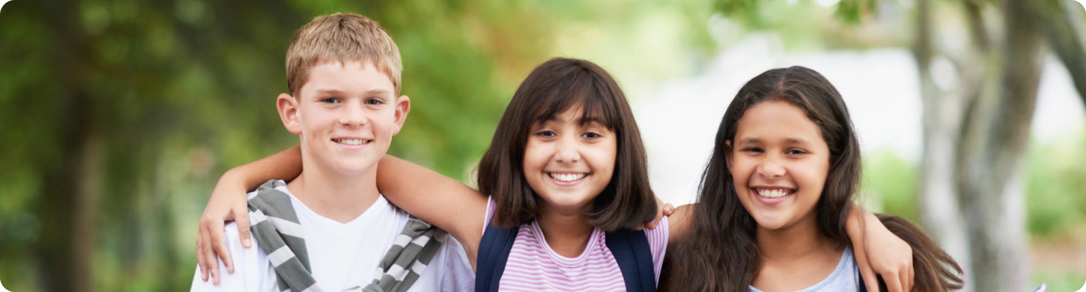 Three smiling children standing outdoors with their arms around each other’s shoulders, with greenery in the blurred background.
