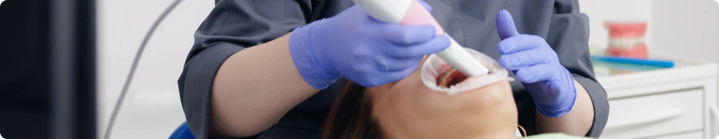 A dentist wearing purple gloves uses a dental scanner inside a patients mouth, who is lying back in a dental chair. Dental models and equipment are visible in the background.